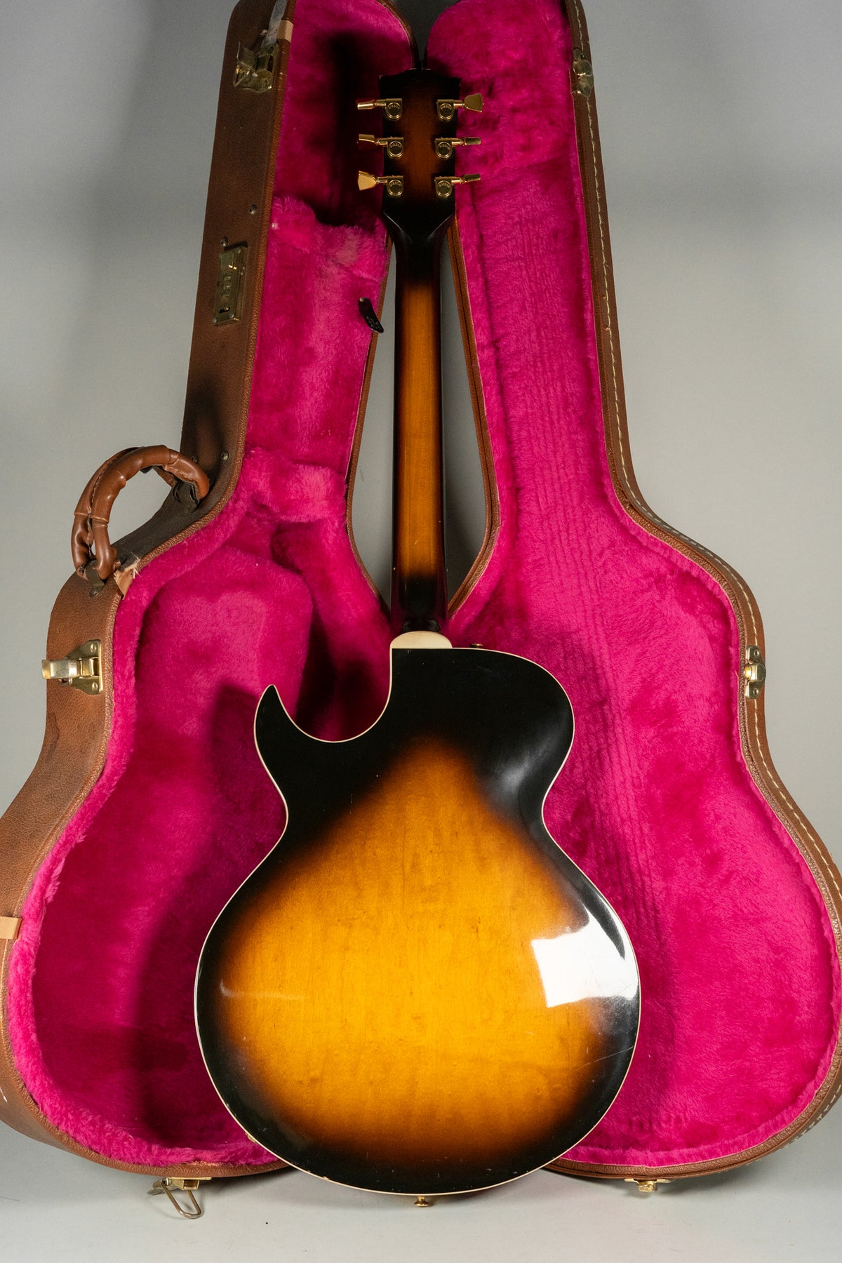 The guitar's sunburst back and neck are visible, with gold tuners and a brown hard case with bright pink plush lining.