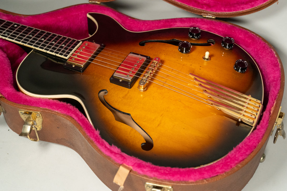 The sunburst hollow-body guitar, with red pickups and a gold trapeze tailpiece, rests in a brown case with pink plush lining.