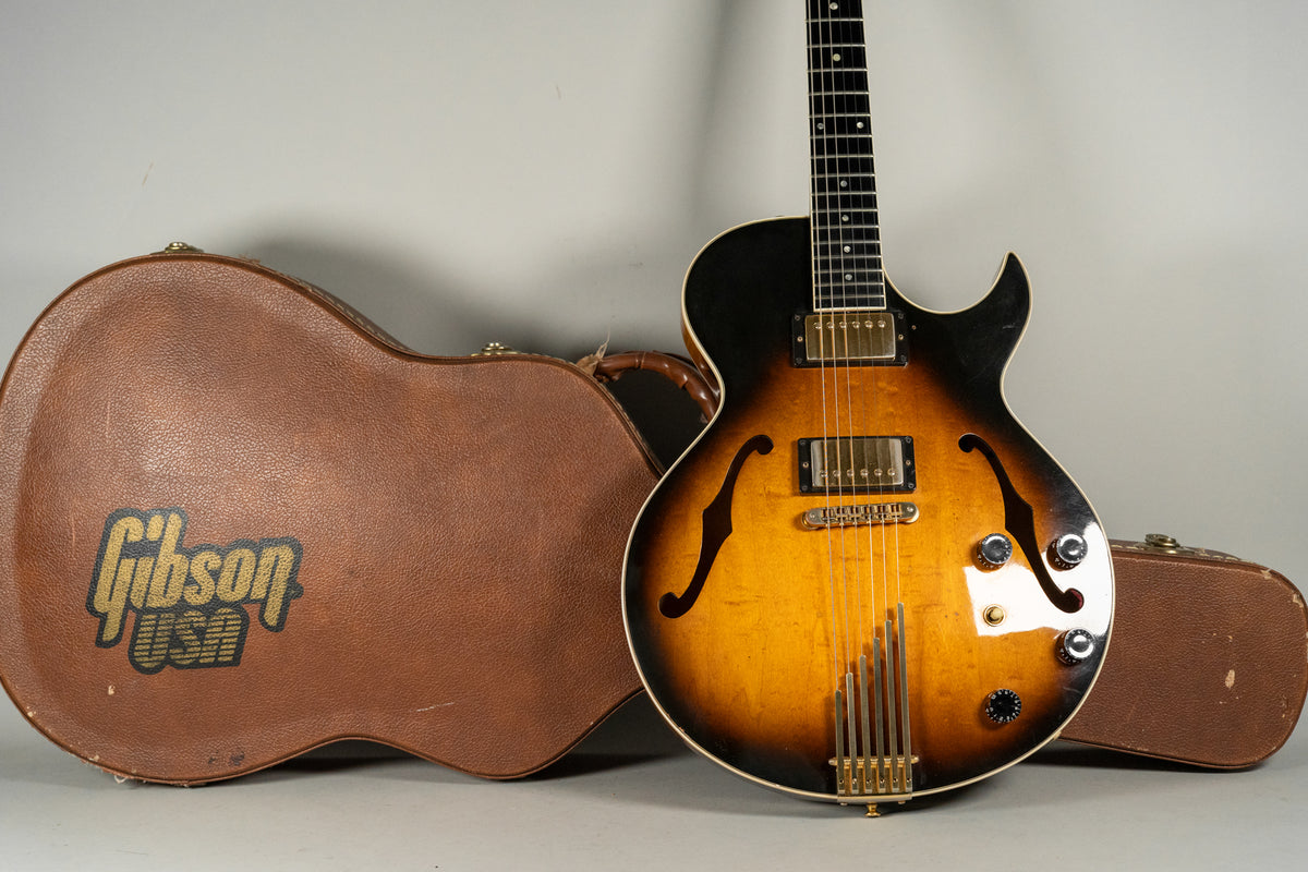 Sunburst archtop guitar with gold hardware, f-holes, and four control knobs, angled next to its brown hardshell case.