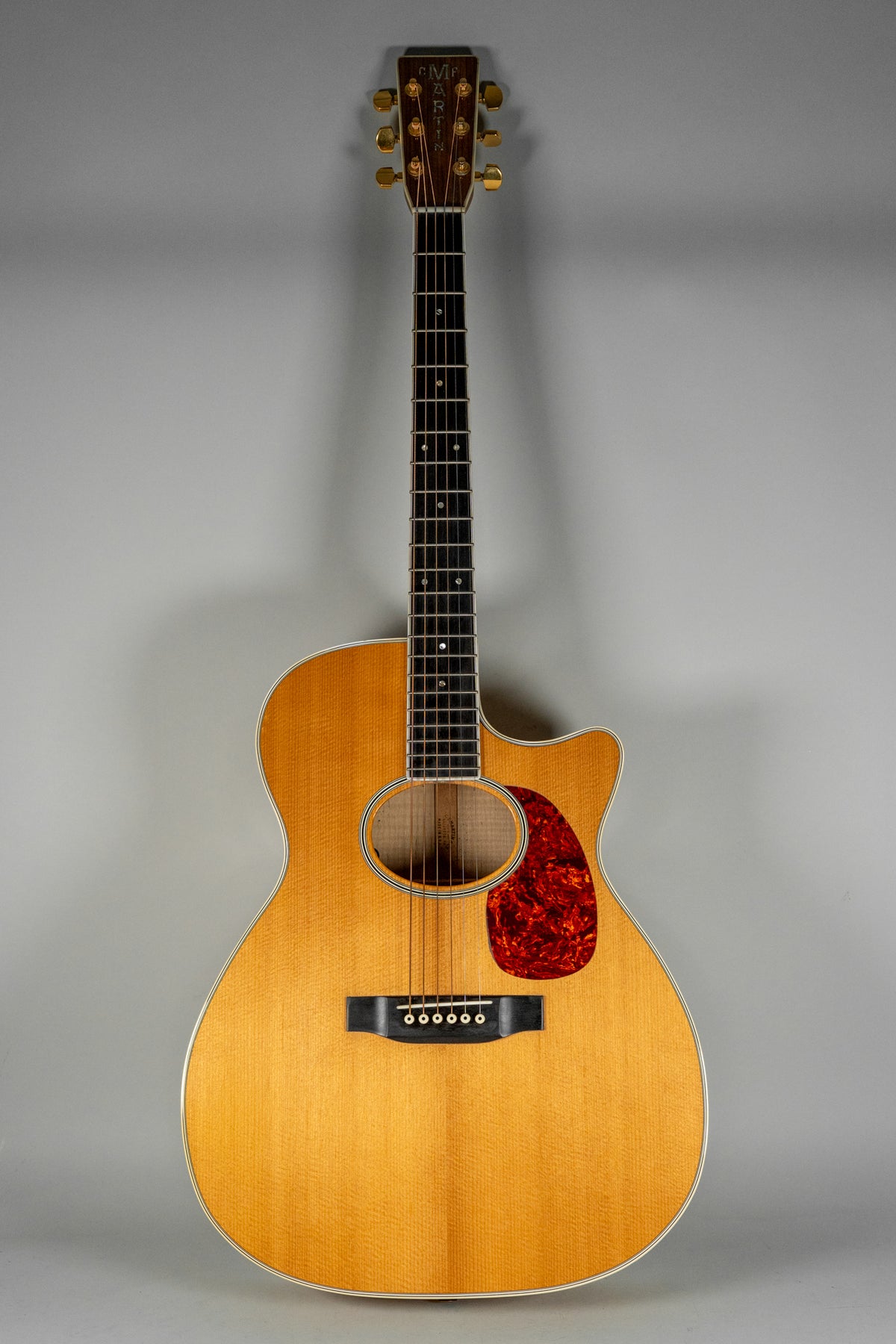 Full-length view of a light-wood acoustic guitar with a cutaway body, red tortoiseshell pickguard, and gold tuners.
