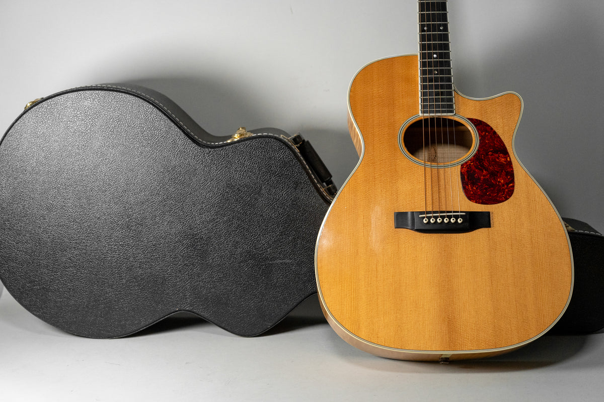 The natural finish acoustic guitar with a cutaway body and tortoiseshell pickguard is shown next to its open black hard case.