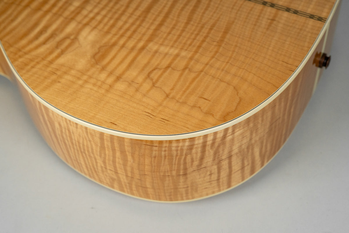 The upper bout and side of a natural flame maple acoustic guitar, showing light wood grain and cream binding.
