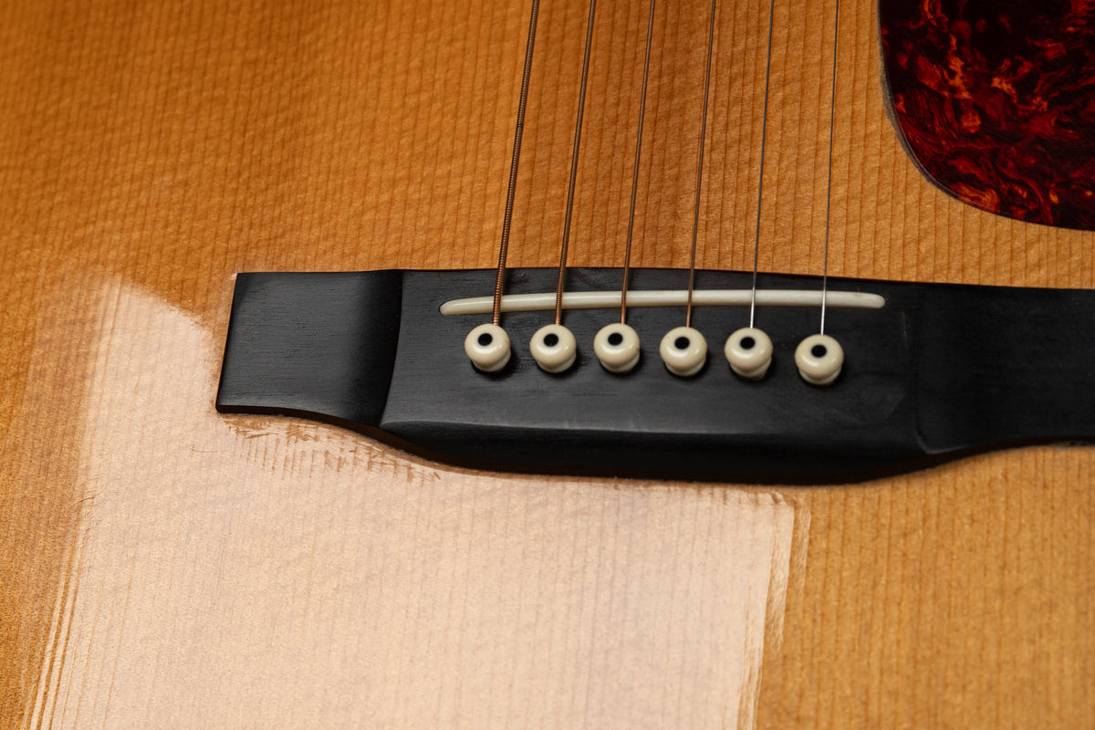 Close-up of the guitar's natural finish body, showing the black bridge, white saddle, and six cream bridge pins, with pick wear below.