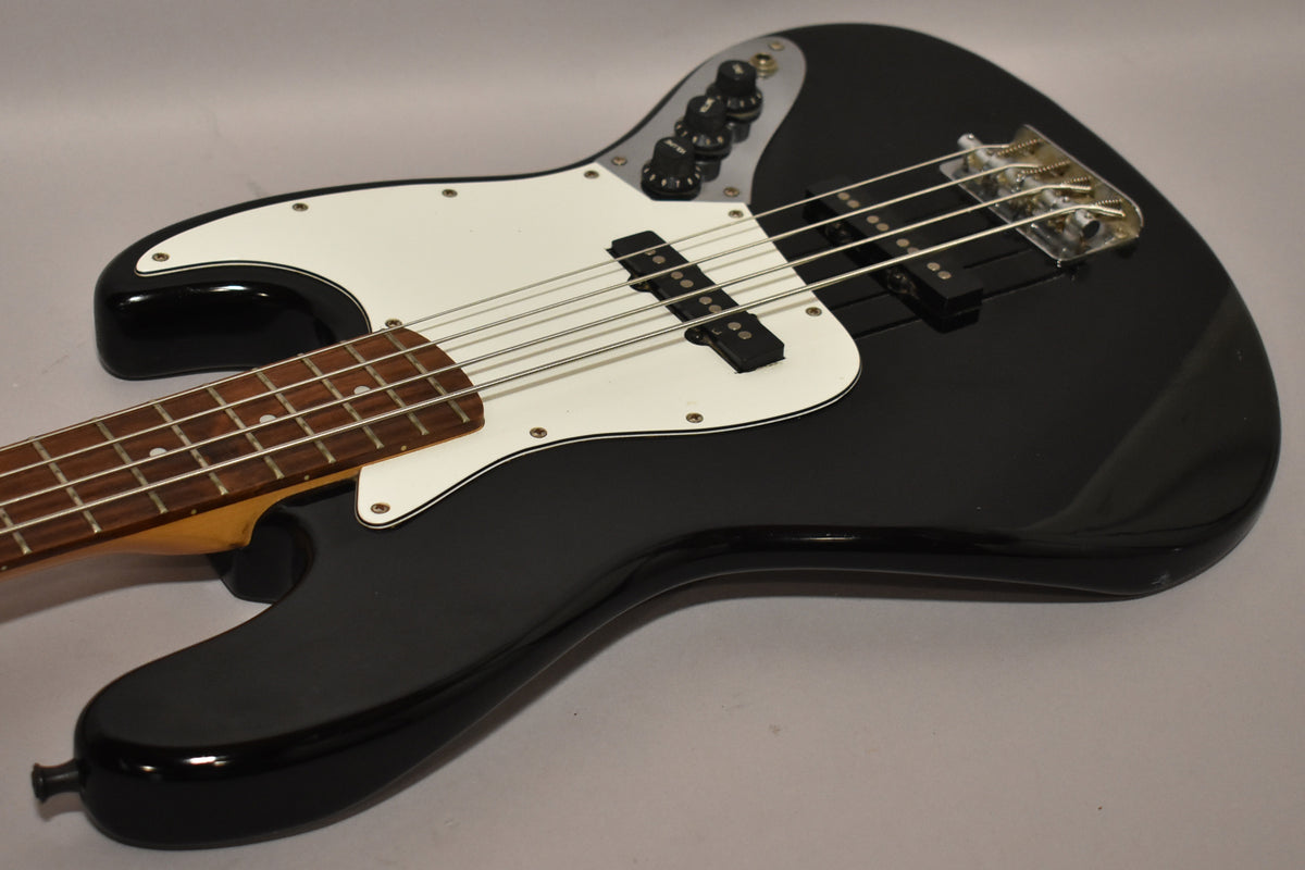 Black Jazz Bass body, angled from the lower bout, showing the white pickguard, two pickups, and control plate with four black knobs.