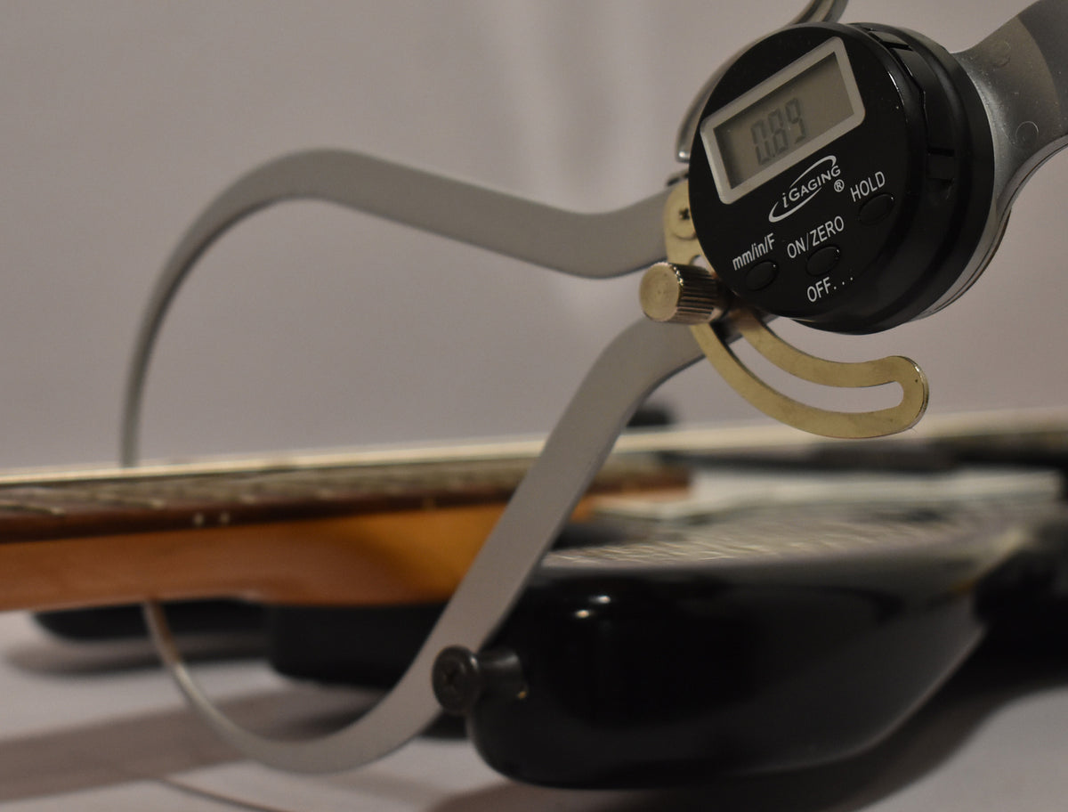 A digital caliper measures the black bass's neck relief, showing 0.89mm, with a worn maple fretboard visible.