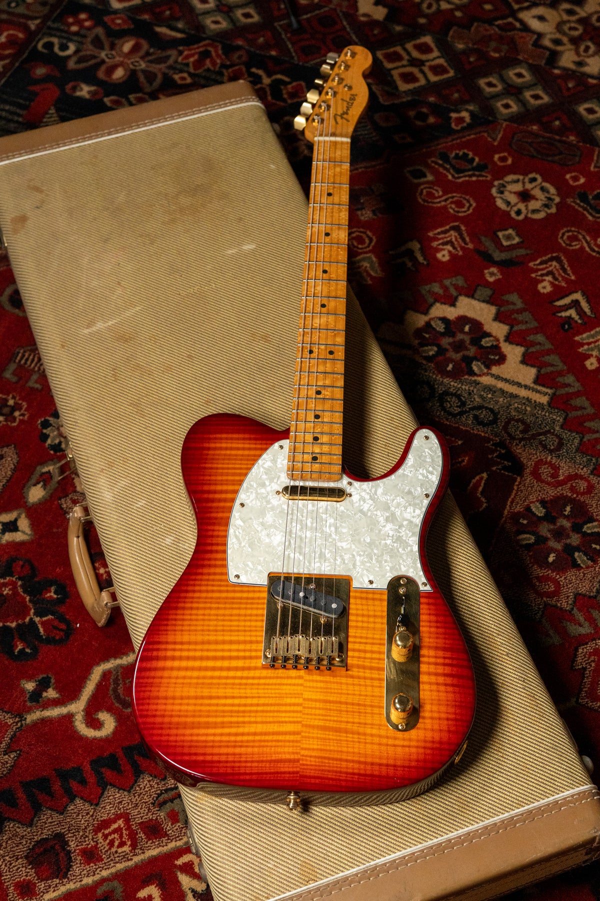 Full view of a sunburst Telecaster with a flame top, pearloid pickguard, gold hardware, and a maple neck, resting on a tweed case.