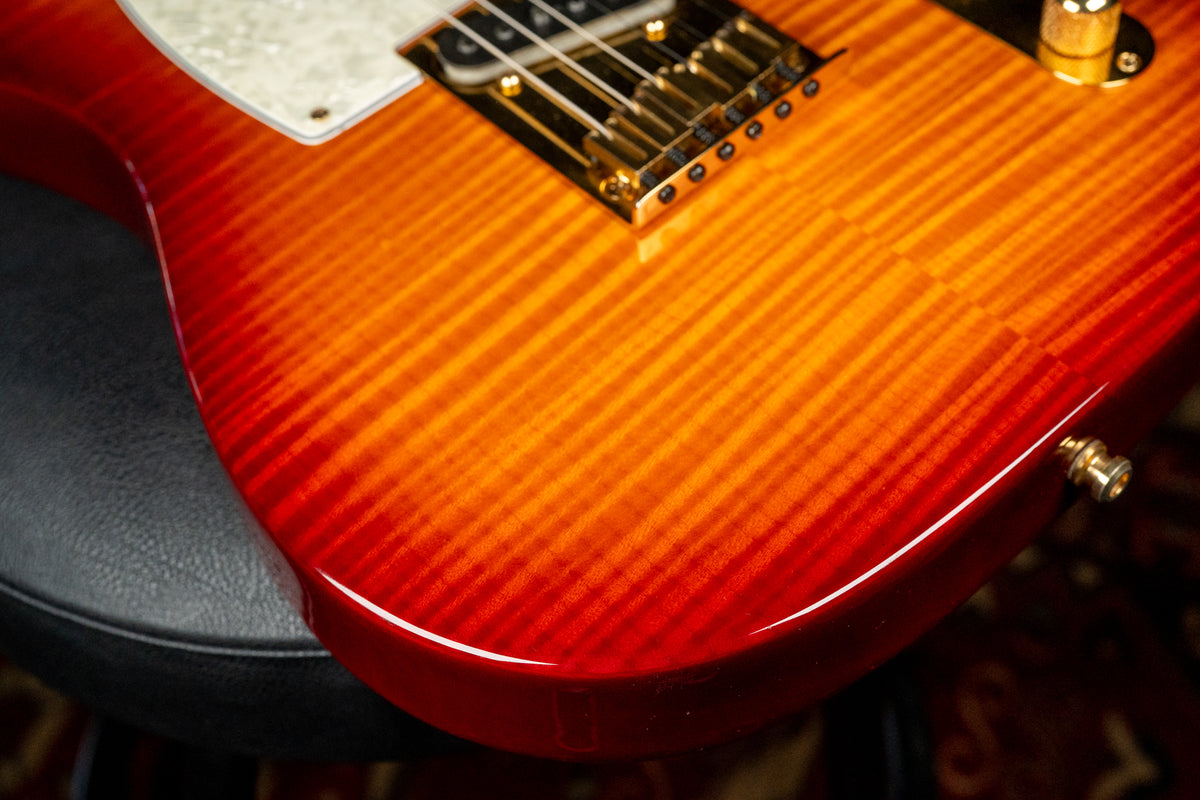 Close-up of the lower body of a glossy sunburst flame-top guitar, showing a gold bridge, white pearloid pickguard, and strap button.