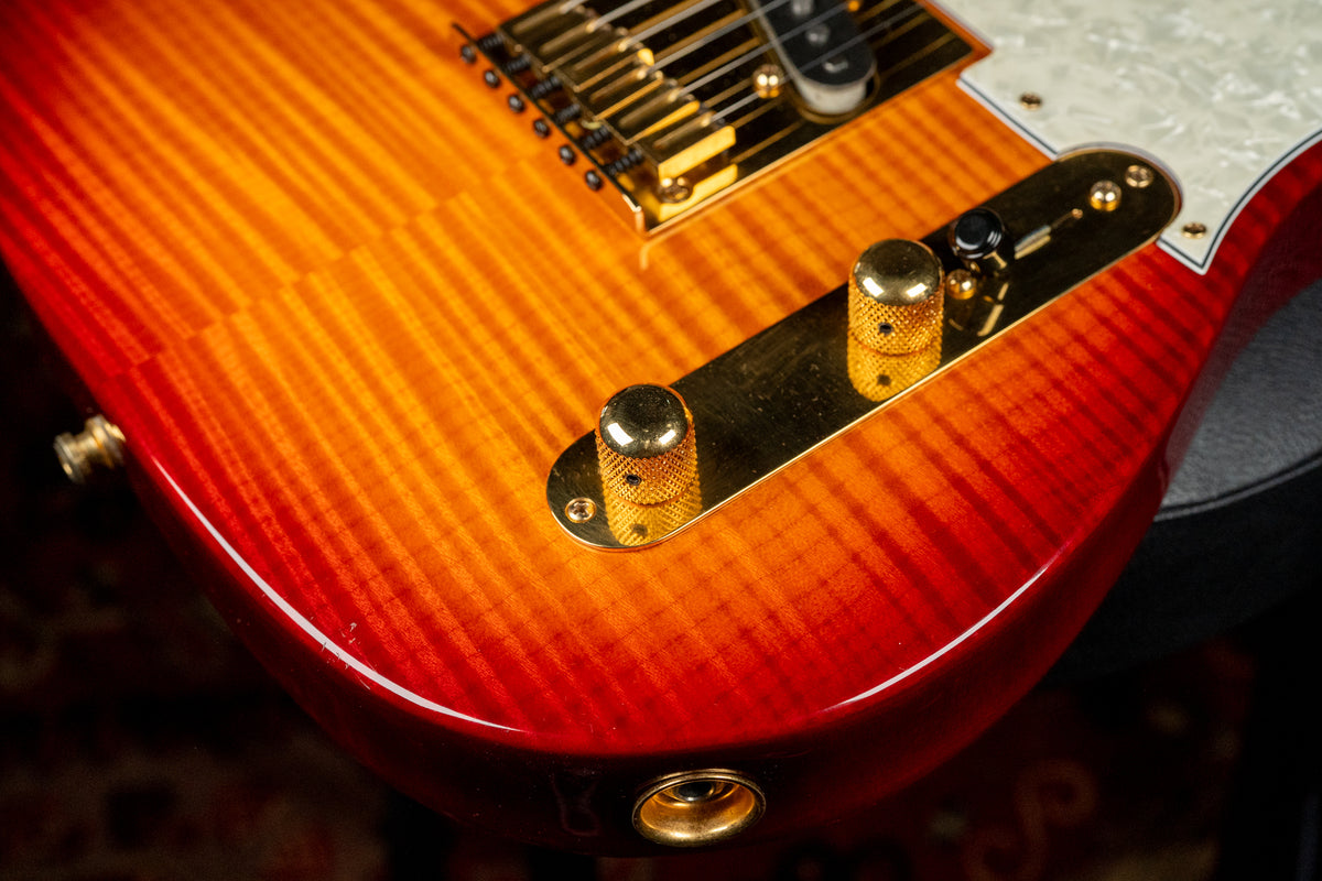 Close-up of a sunburst flame top Telecaster body, showing gold hardware, two knurled gold control knobs, and a small chip on the lower bout.