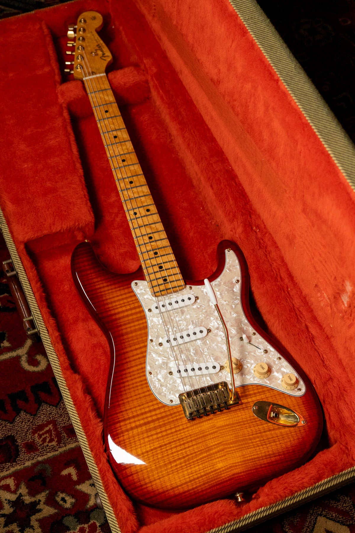 A Stratocaster in a tweed case with red plush lining, showing its flamed maple sunburst finish, pearloid pickguard, and gold hardware.