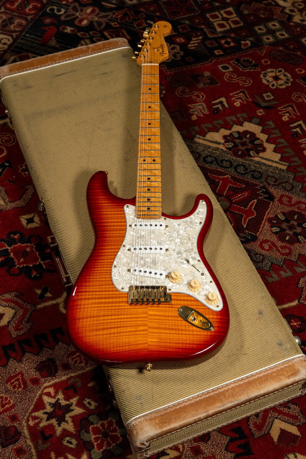 A Stratocaster with a cherry sunburst flame top, white pearloid pickguard, gold hardware, and a maple neck, resting on a tweed case.