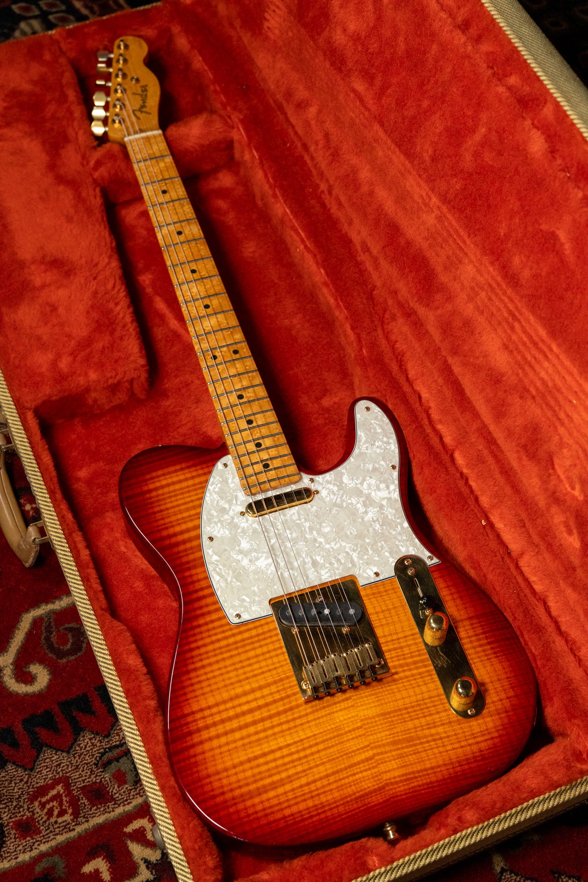 A Telecaster with a flamed sunburst finish, gold hardware, and a pearloid pickguard rests in a red velvet case.