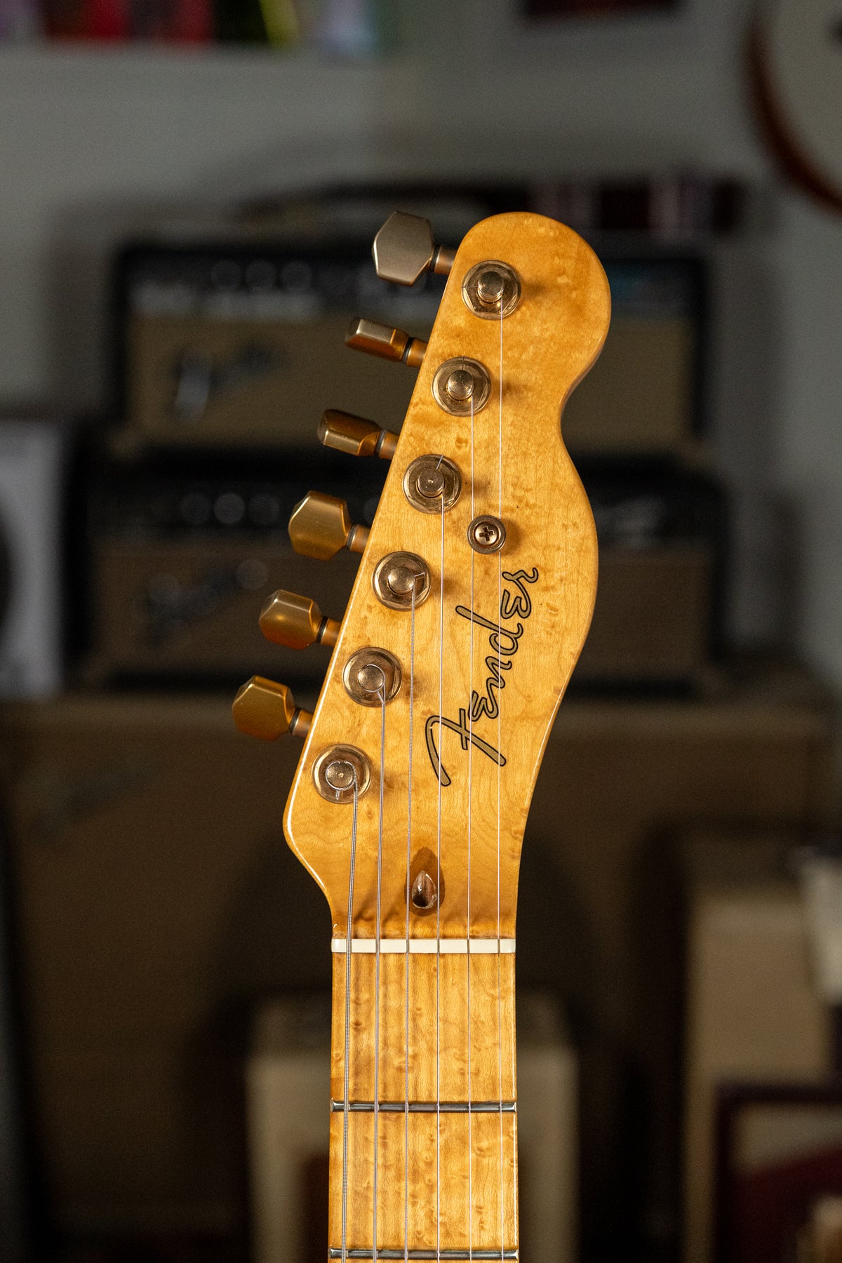 Close-up of a light brown figured maple headstock with six gold tuners and a matching maple fretboard with light strings.