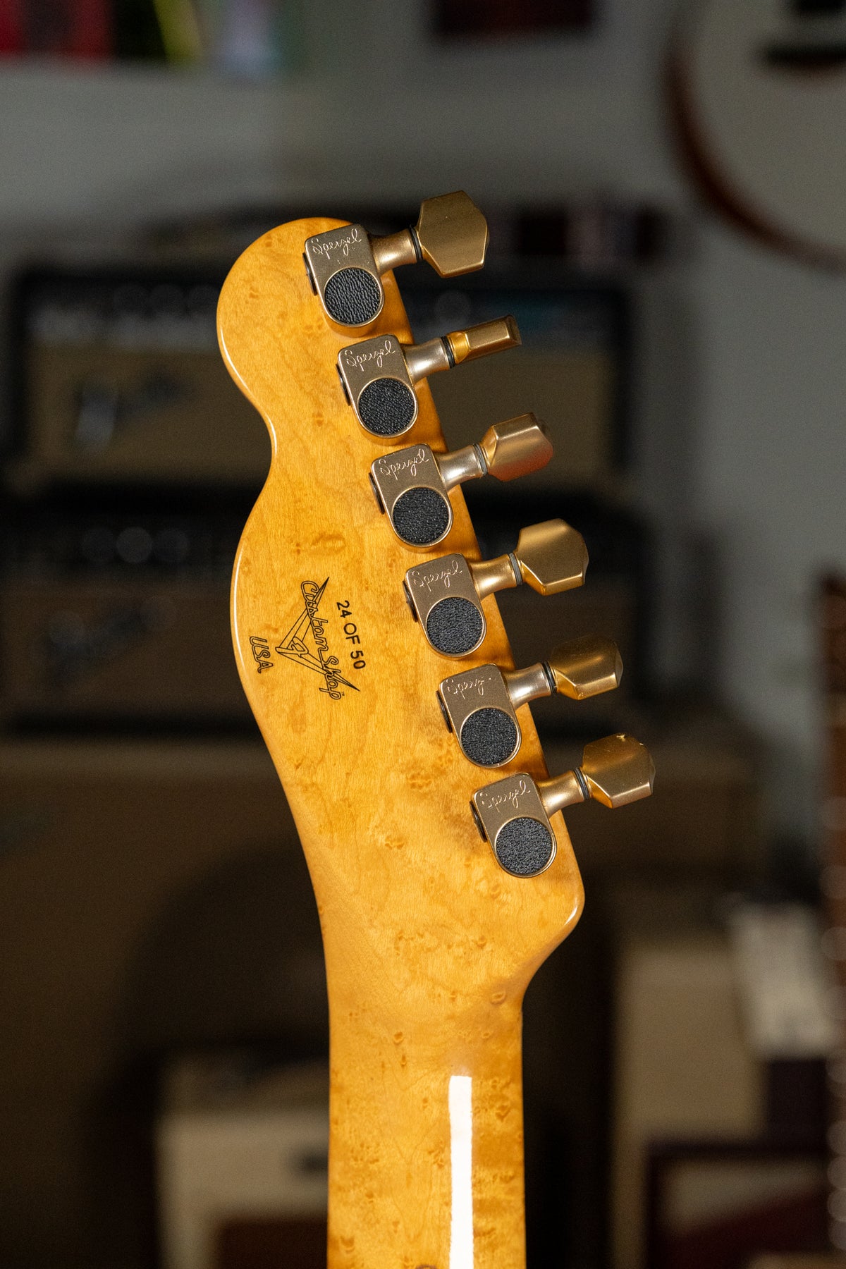 The back of a light-colored, burled wood headstock with six gold tuners, angled slightly to the left.