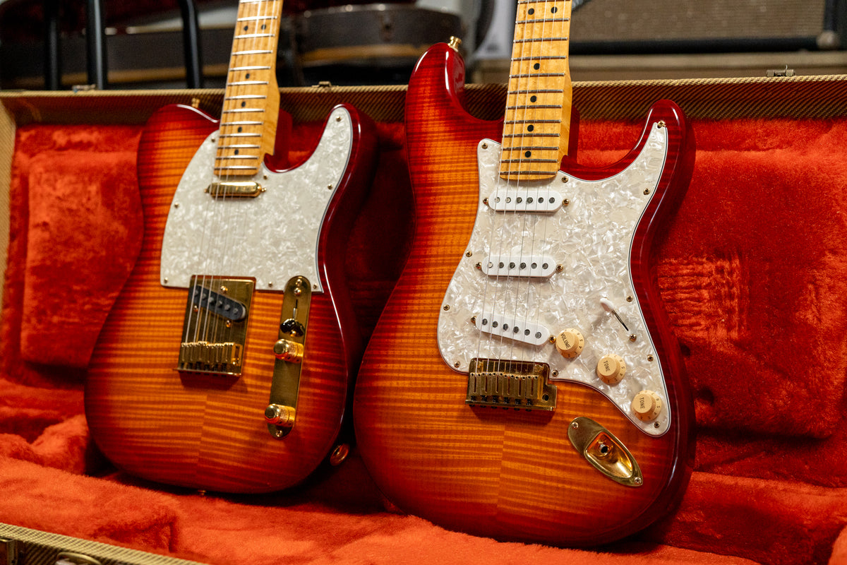 Two flame-top guitars with cherry sunburst finish, gold hardware, and pearloid pickguards rest in a red velvet-lined hard case.