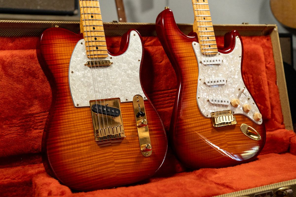 Two guitars with amber-to-red sunburst finishes and pearloid pickguards rest in a red velvet case, showing their bodies and necks.