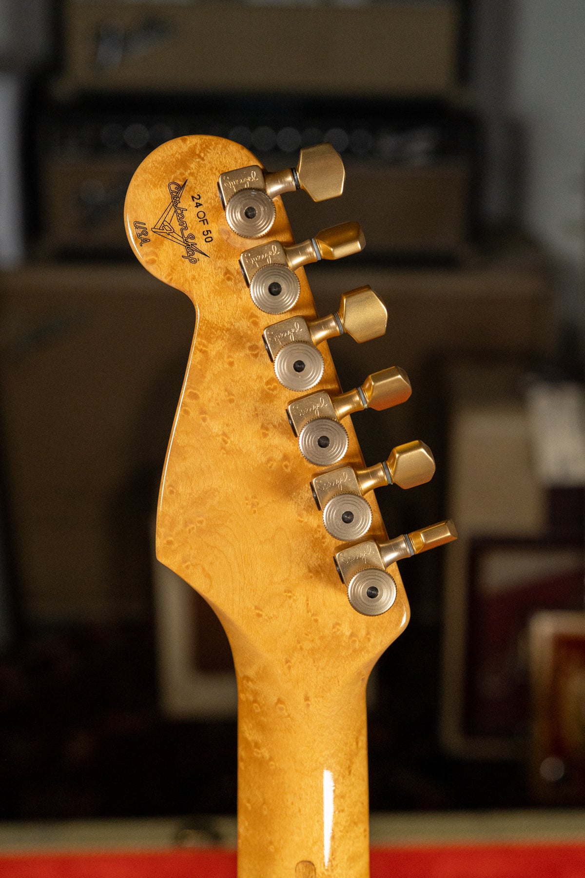 The guitar's headstock and upper neck, made of light-colored wood with a bird's-eye maple grain, are angled to the right, showing six gold tuners.