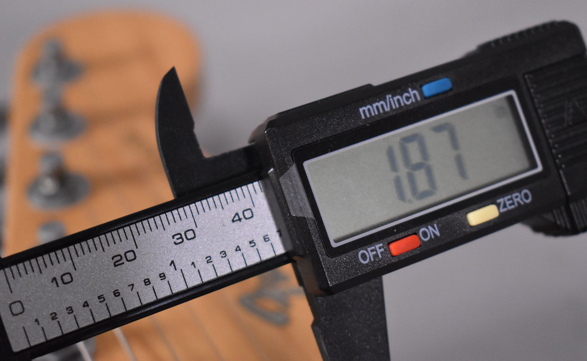 A digital caliper measures the nut width of the blonde Stratocaster's headstock, displaying "1.87" on its screen.