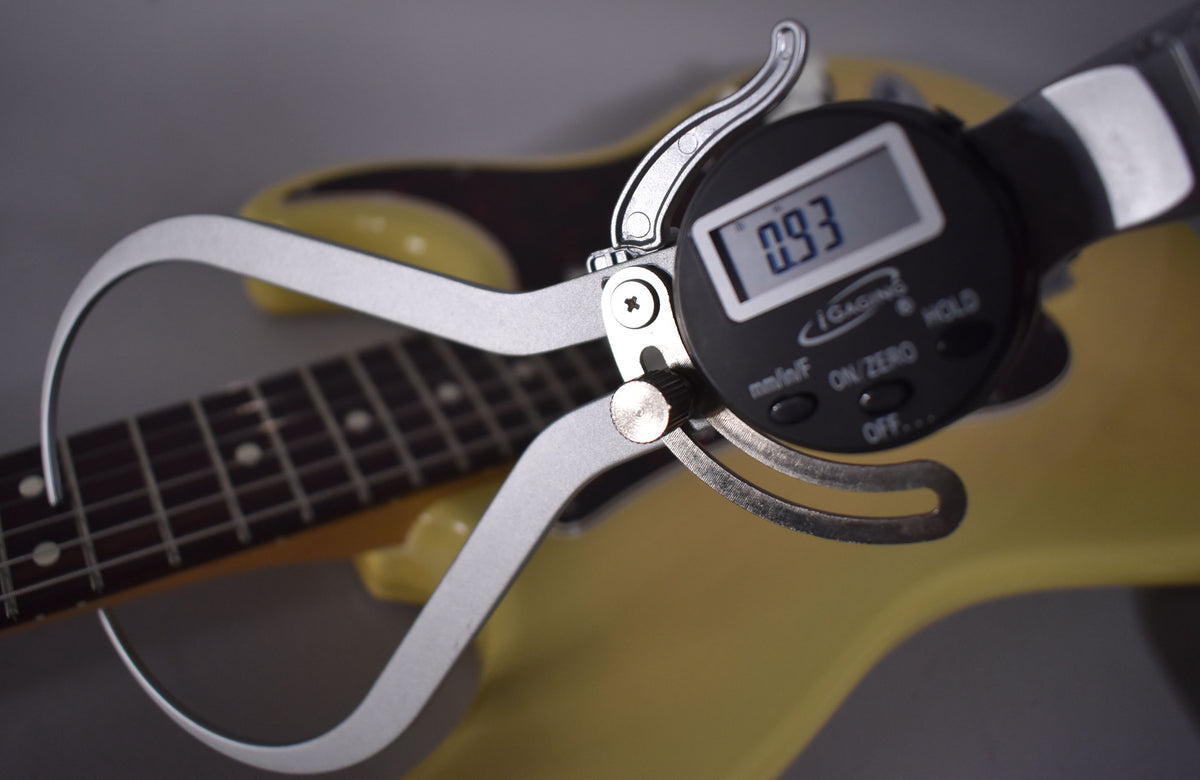A digital caliper measures the fret height on the dark fretboard of a vintage blonde electric guitar.