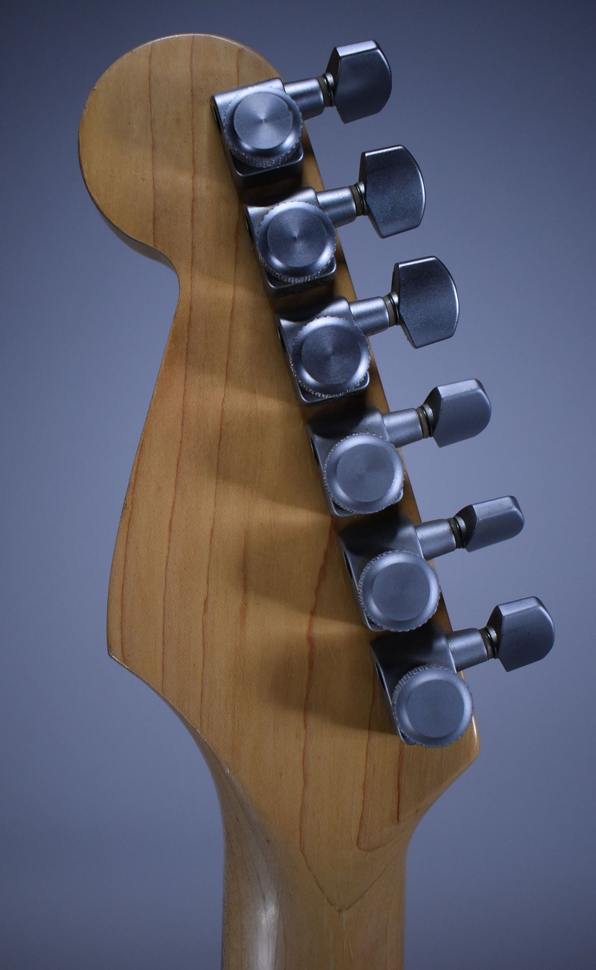 The back of a light blonde guitar headstock and neck, showing six brushed chrome tuners with some light wear.
