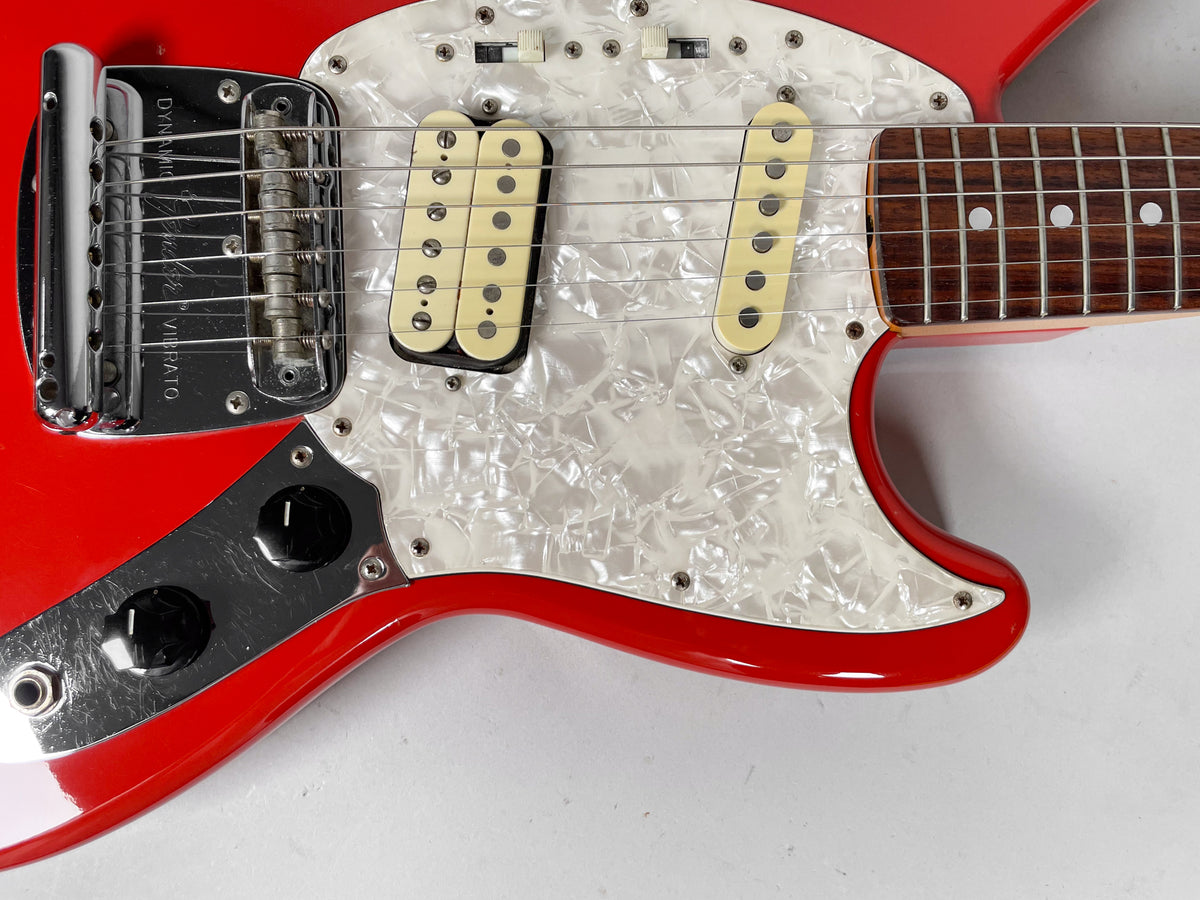 Close-up of a red guitar body with a pearloid pickguard, chrome vibrato, two cream pickups, and two black control knobs.