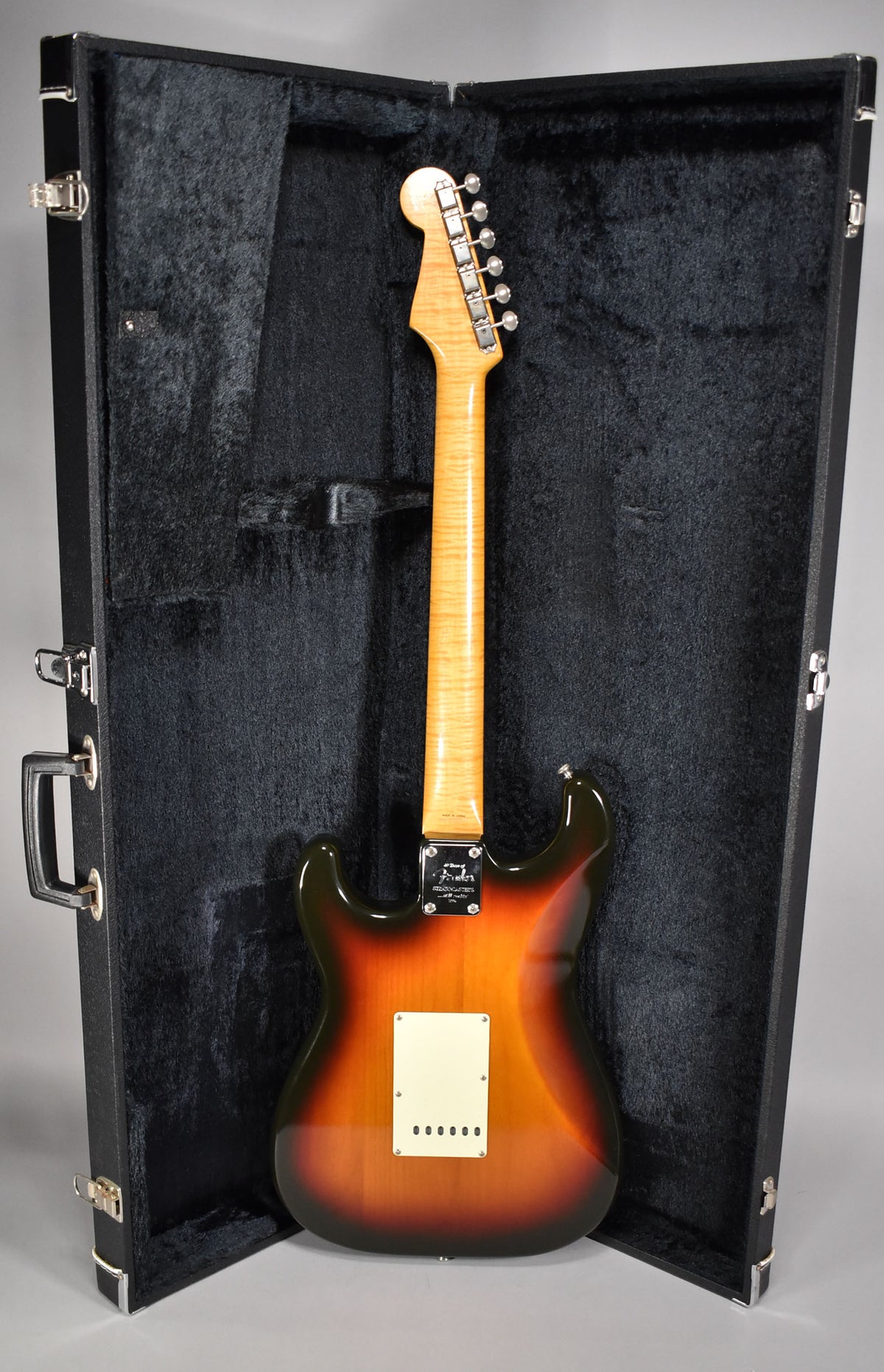 The guitar's back, in a glossy three-tone sunburst, is shown in its open black hard case, revealing a light maple neck and headstock.