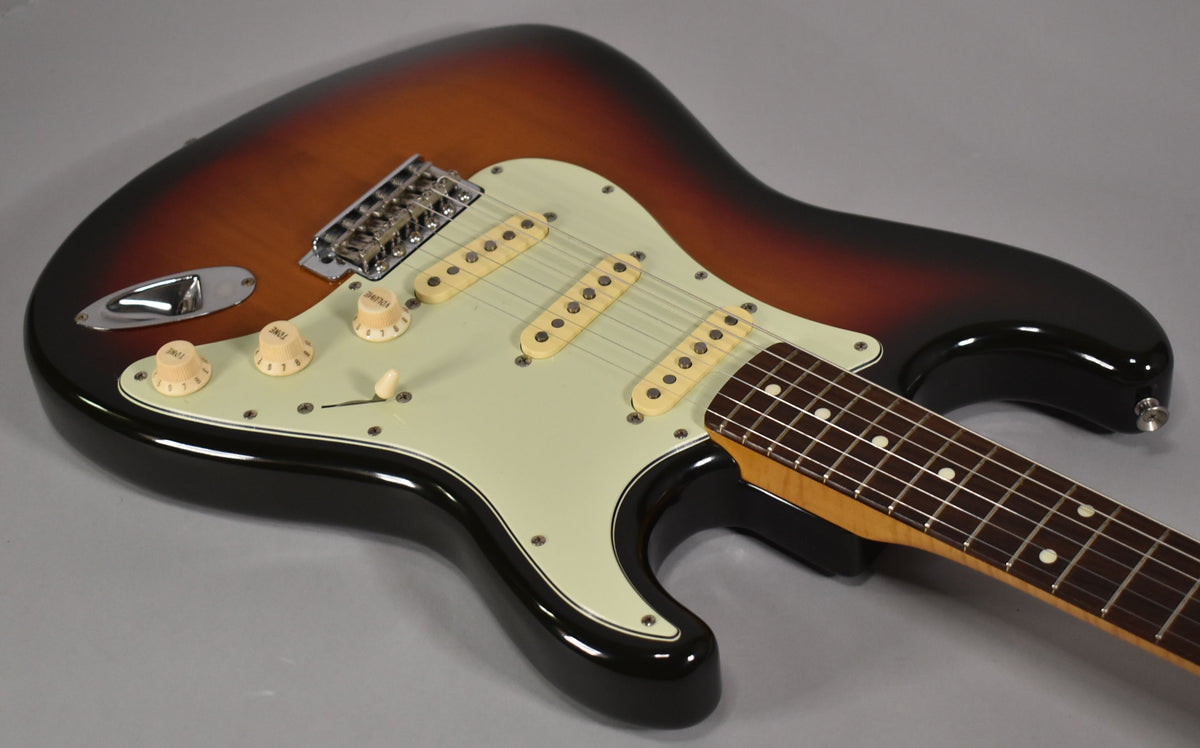 Sunburst electric guitar, angled from above, showing the body, pickguard, three single-coil pickups, and part of the neck.