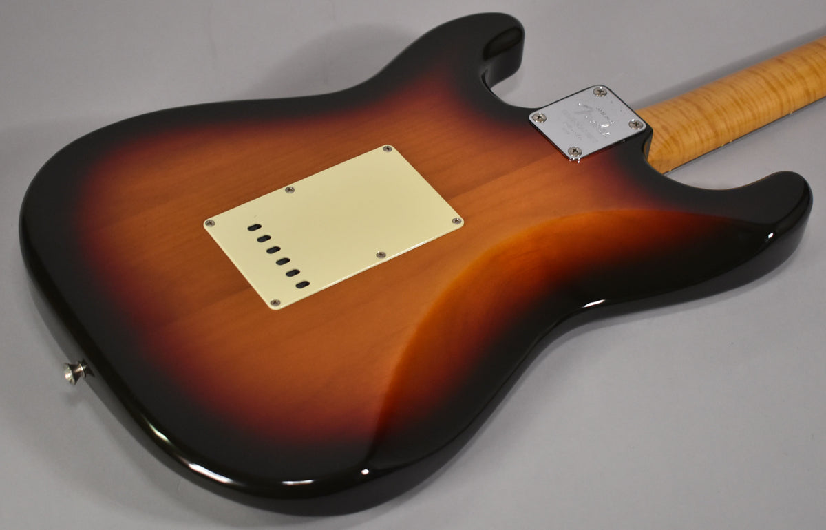 The guitar's glossy three-tone sunburst body is seen from the back, revealing a cream tremolo cover and a flamed maple neck.