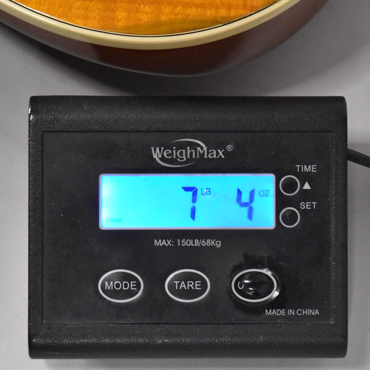 The guitar's amber body is visible above a black digital scale displaying "7 LB 4 OZ".