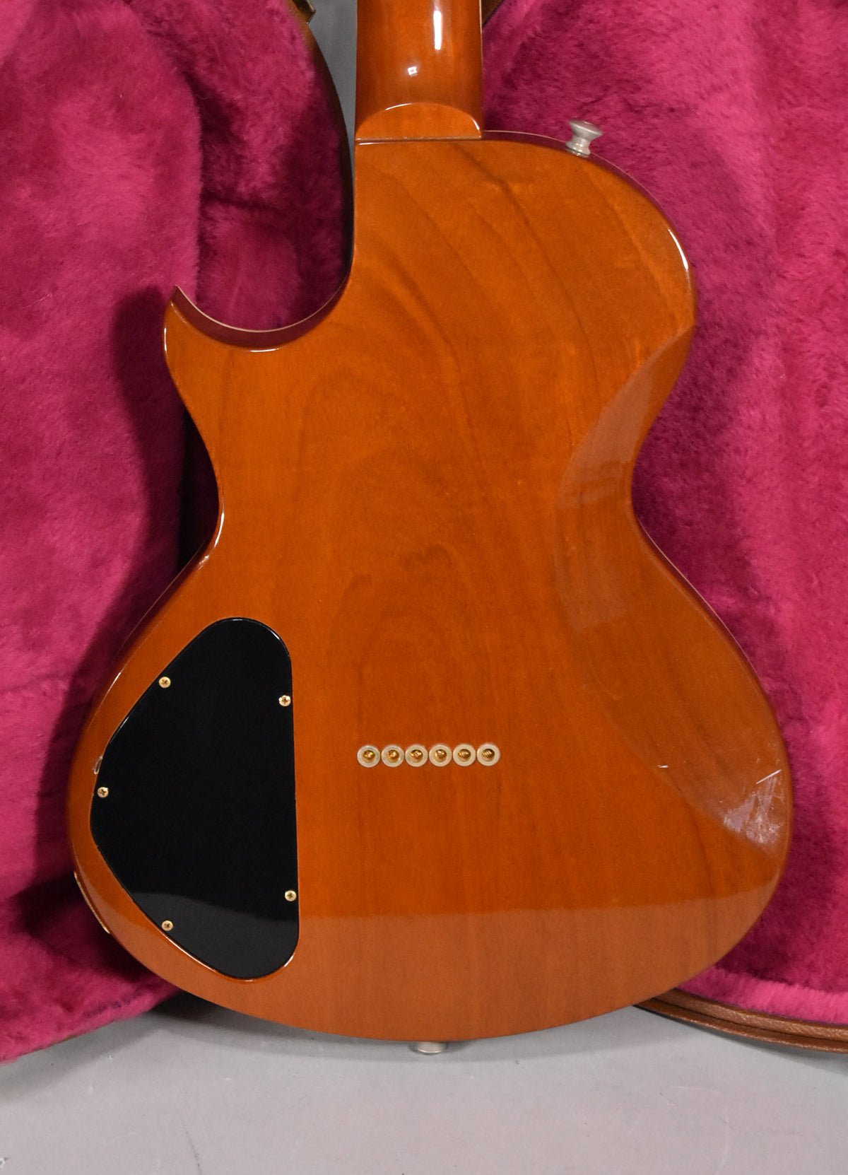 The back of a transparent amber guitar shows a black control plate, six gold string ferrules, and light scratches on the lower bout.