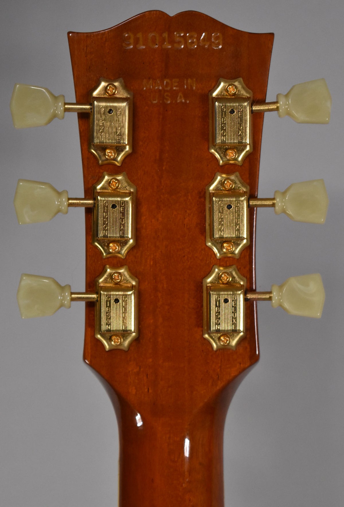 The guitar's headstock, viewed from the back, shows six gold-plated tuners with cream plastic buttons on a glossy amber finish.