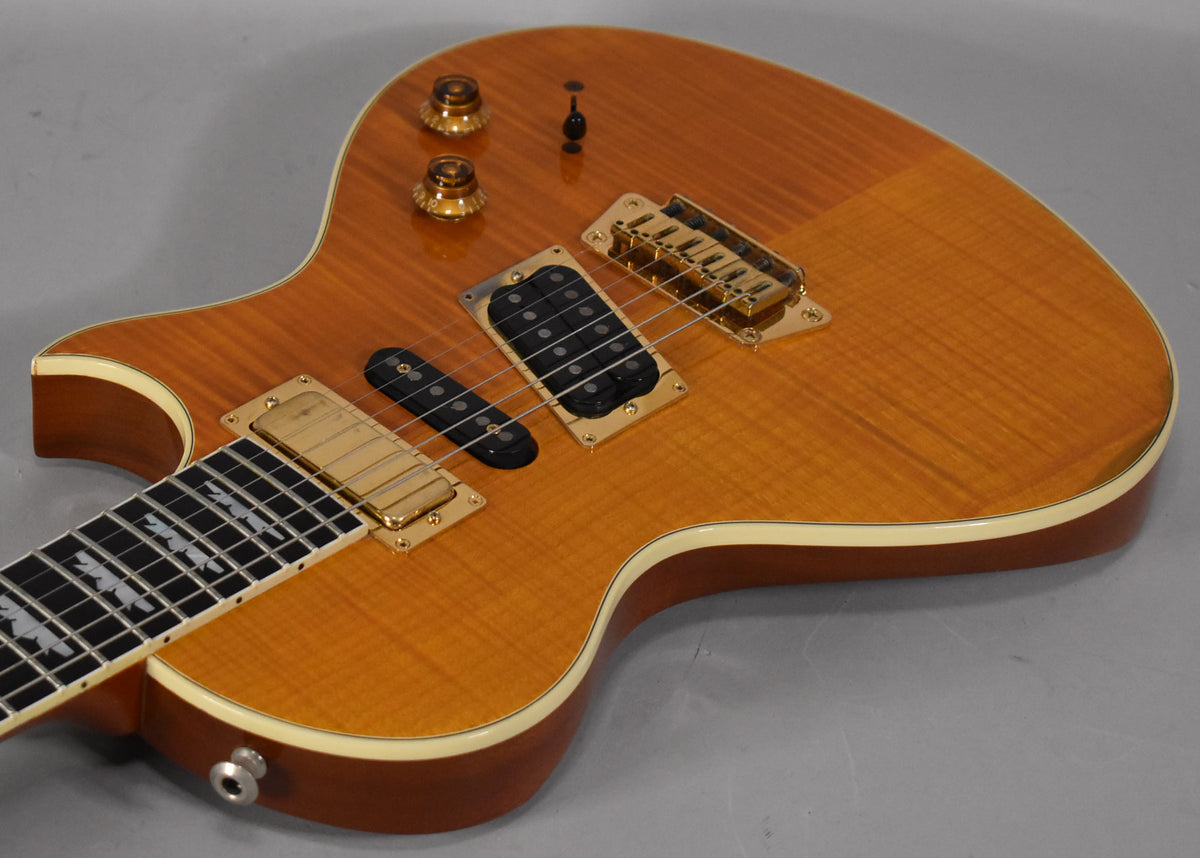The amber flamed maple guitar body, with cream binding, shows gold hardware and two humbuckers, one with a black cover.