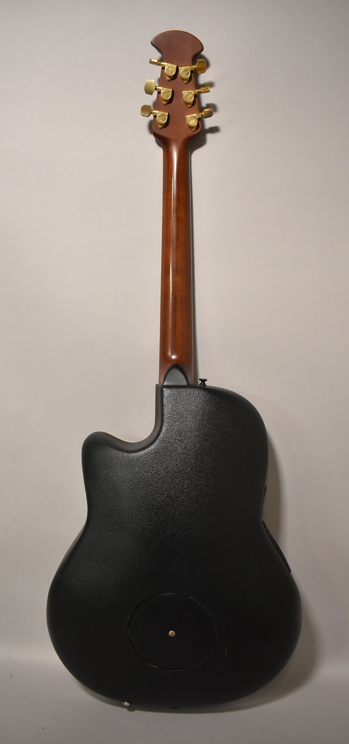 Rear view of the electro-acoustic guitar, showing its dark brown neck, gold tuners, and textured black bowl-back body.
