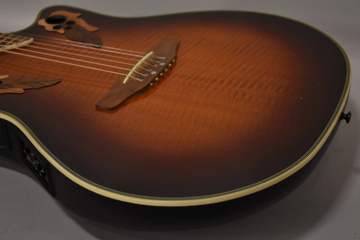 The sunburst electro-acoustic guitar's upper body, showing the dark brown to amber flame maple top, bridge, and leaf-shaped soundholes.