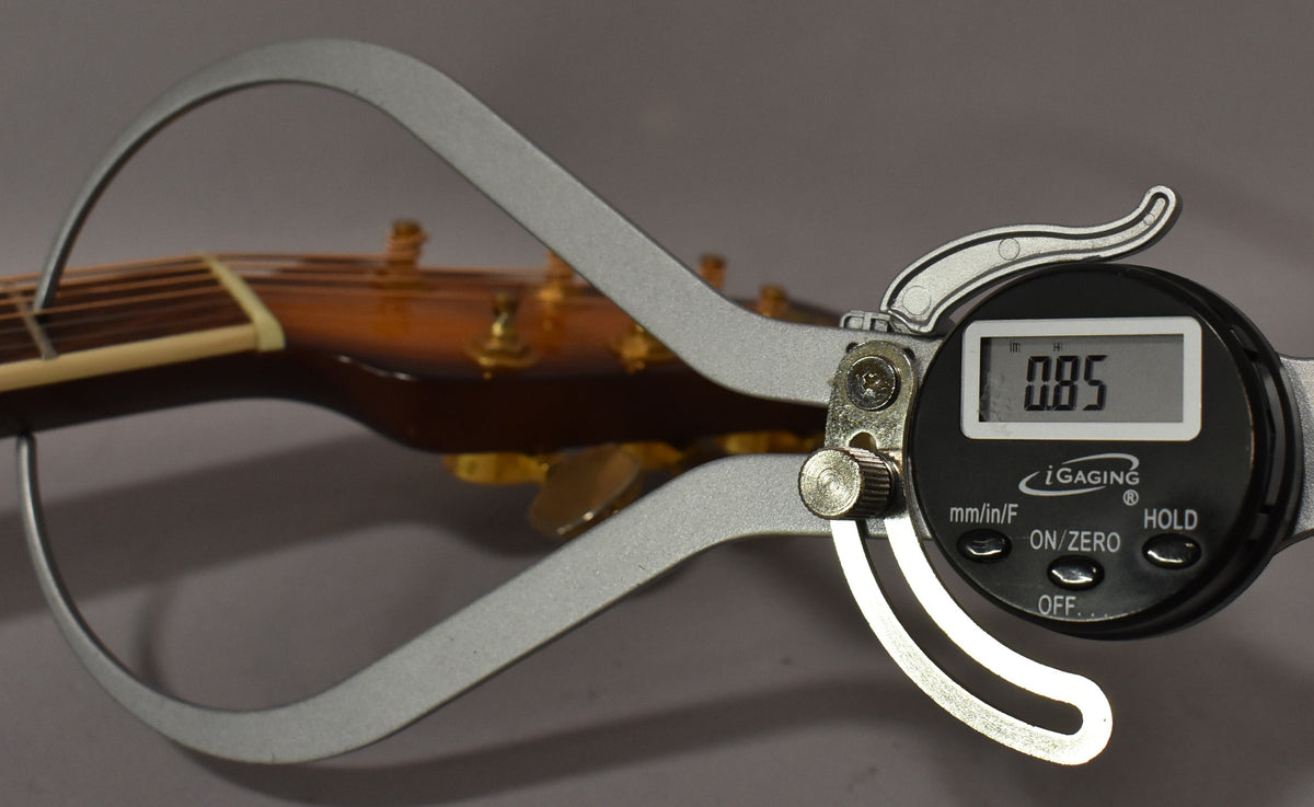 A digital caliper measures the guitar's nut, showing 0.85 on its screen, with the headstock and tuning pegs blurred behind.