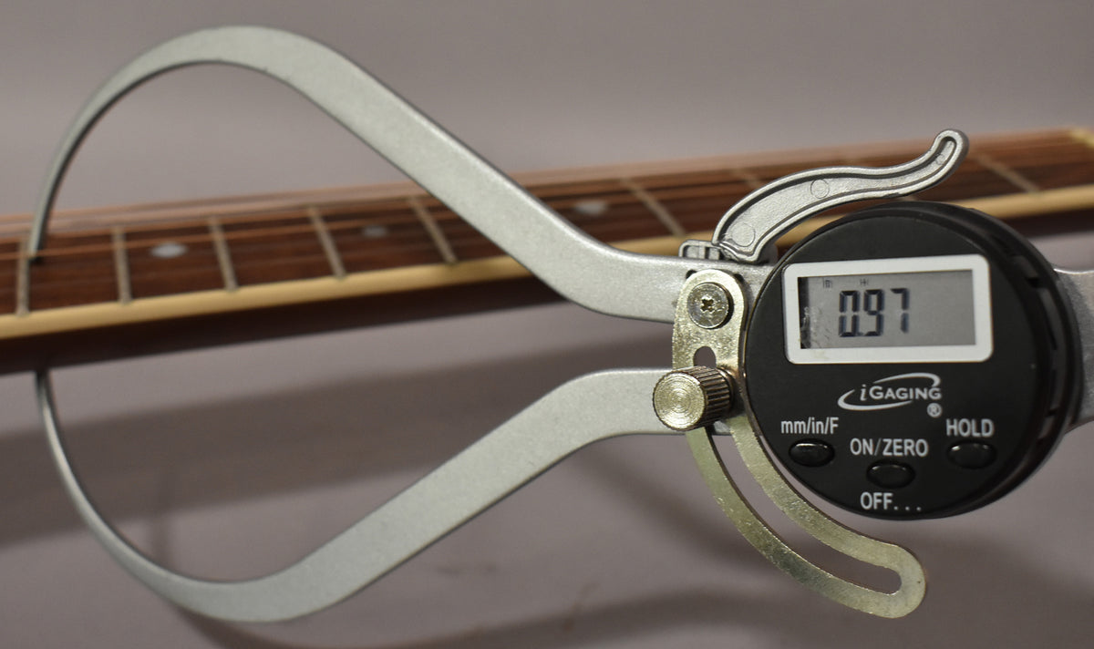 A digital caliper measures the guitar's fretboard, showing a reading of 0.97, with light wear visible on the frets.