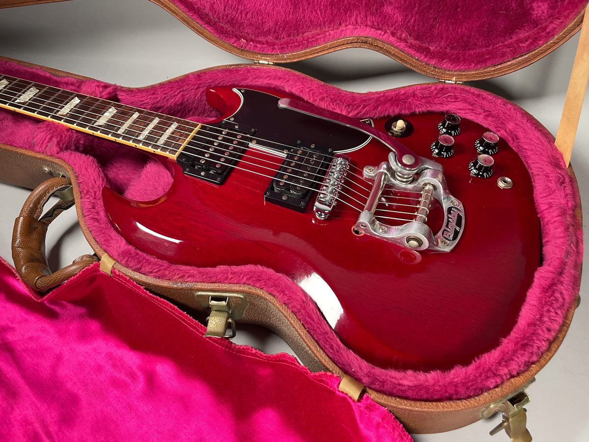 Cherry red SG with a Bigsby vibrato, black pickguard, and four black control knobs, resting in a pink-lined brown case.