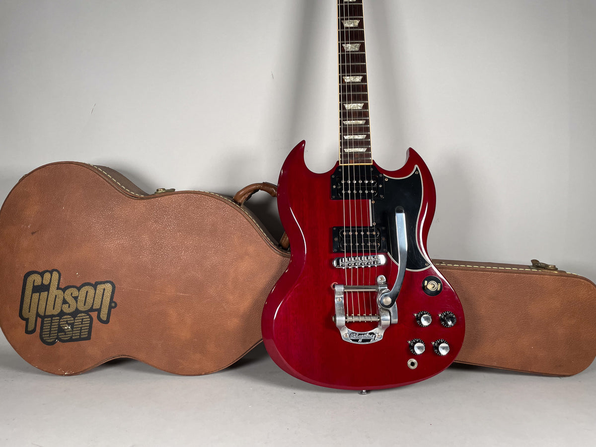 A cherry red SG with a Bigsby vibrato and black pickguard stands next to a brown hardshell case.