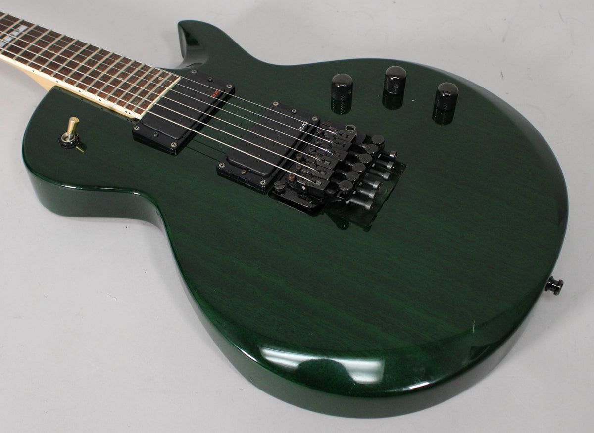 Angled view of a dark green electric guitar with a Floyd Rose tremolo, two humbuckers, three control knobs, and a gold toggle switch.