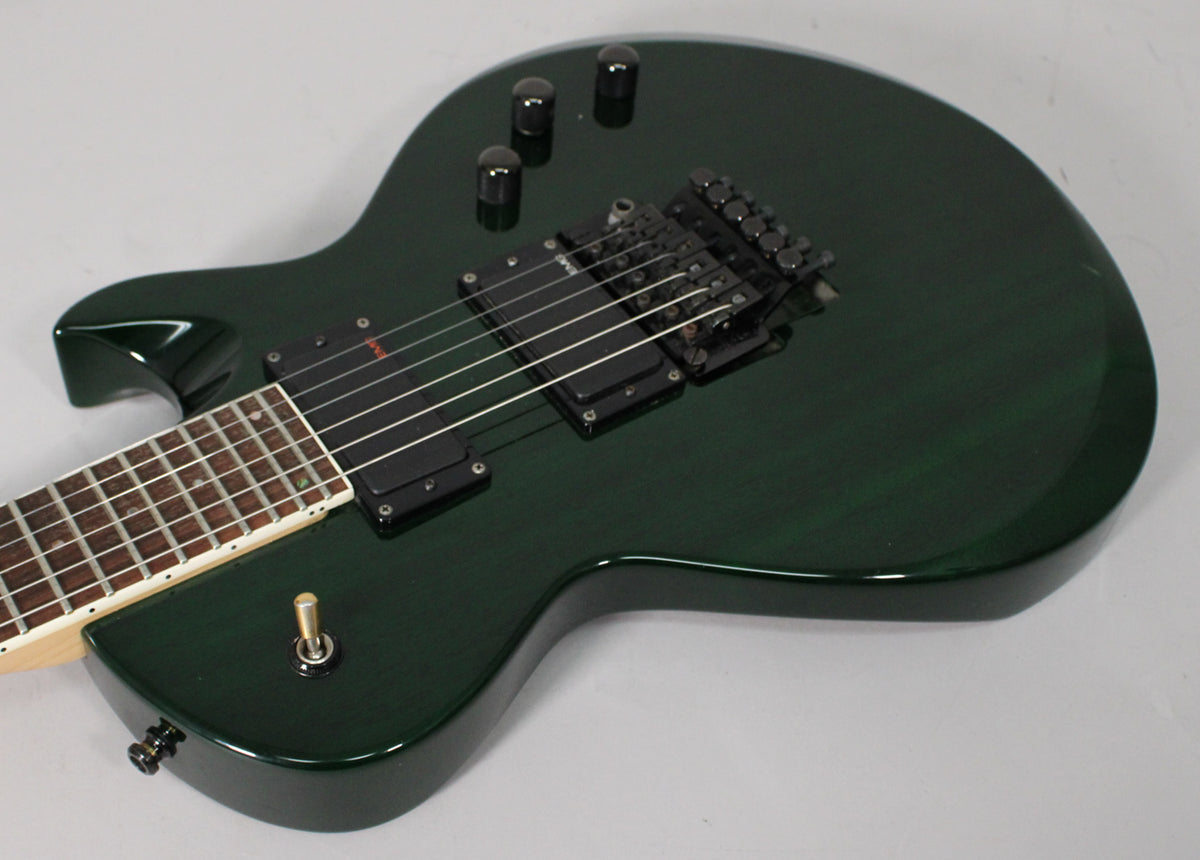 Dark green electric guitar, angled from above, showing the body, black humbuckers, Floyd Rose bridge, and part of the fretboard.