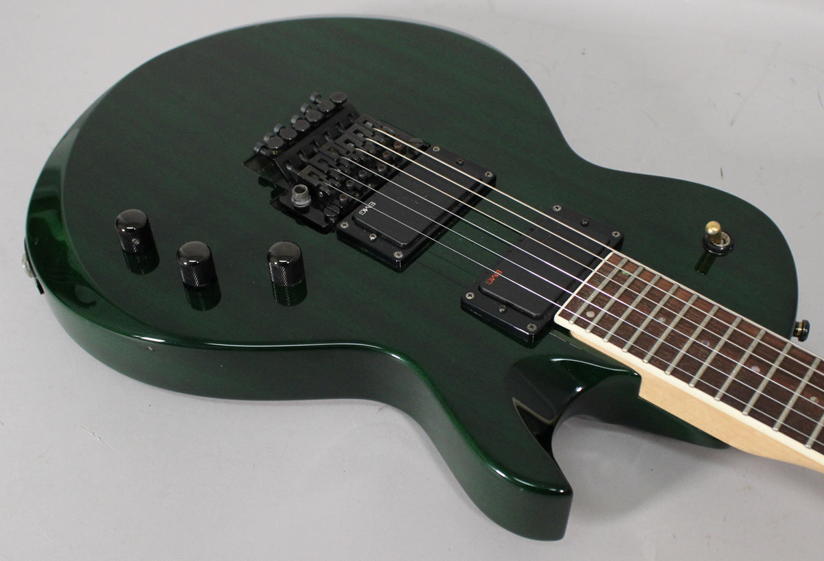 Close-up of a dark green electric guitar, showing the upper body, black Floyd Rose tremolo, two EMG humbuckers, and three black control knobs.