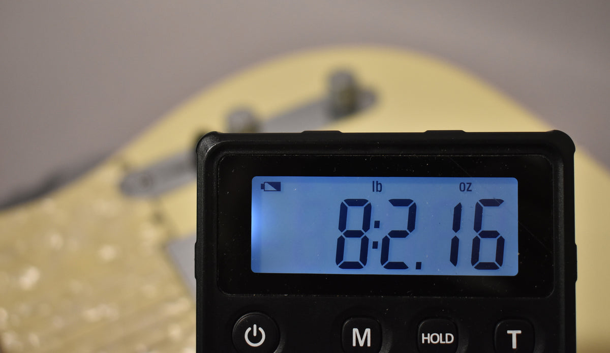 A digital scale displays "8:2.16 lb oz" with a blurry Olympic White Telecaster body in the background.