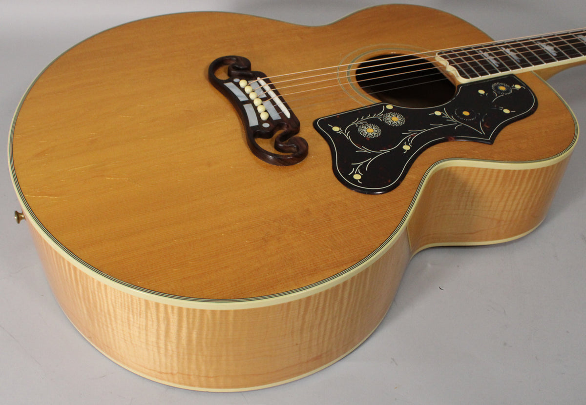 A natural finish acoustic guitar, angled from the lower bout, showing the ornate pickguard, bridge, and flame maple sides.
