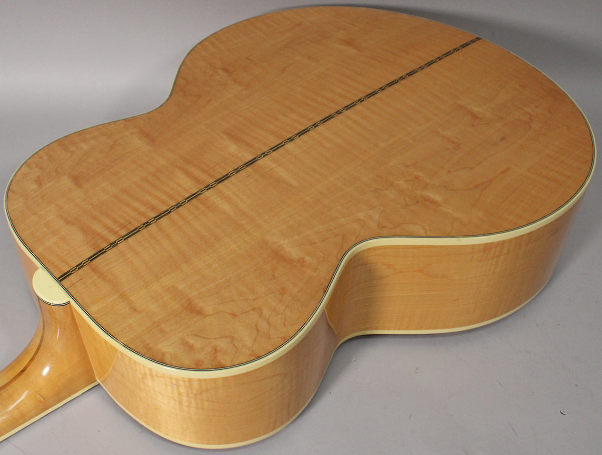 The back of a natural finish acoustic guitar, showing the flame maple grain, a dark center strip, and cream binding.