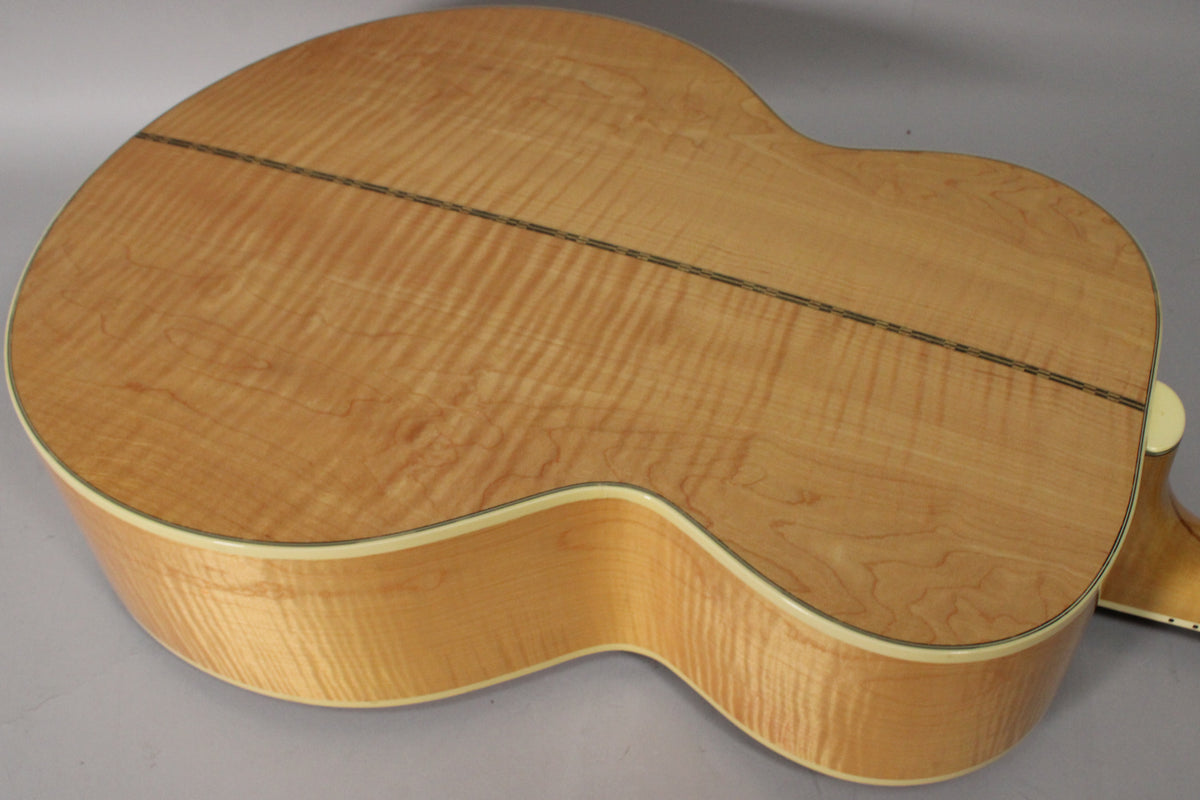 The back of the natural finish acoustic guitar, angled from the upper bout, shows a flame maple grain with a dark center strip.