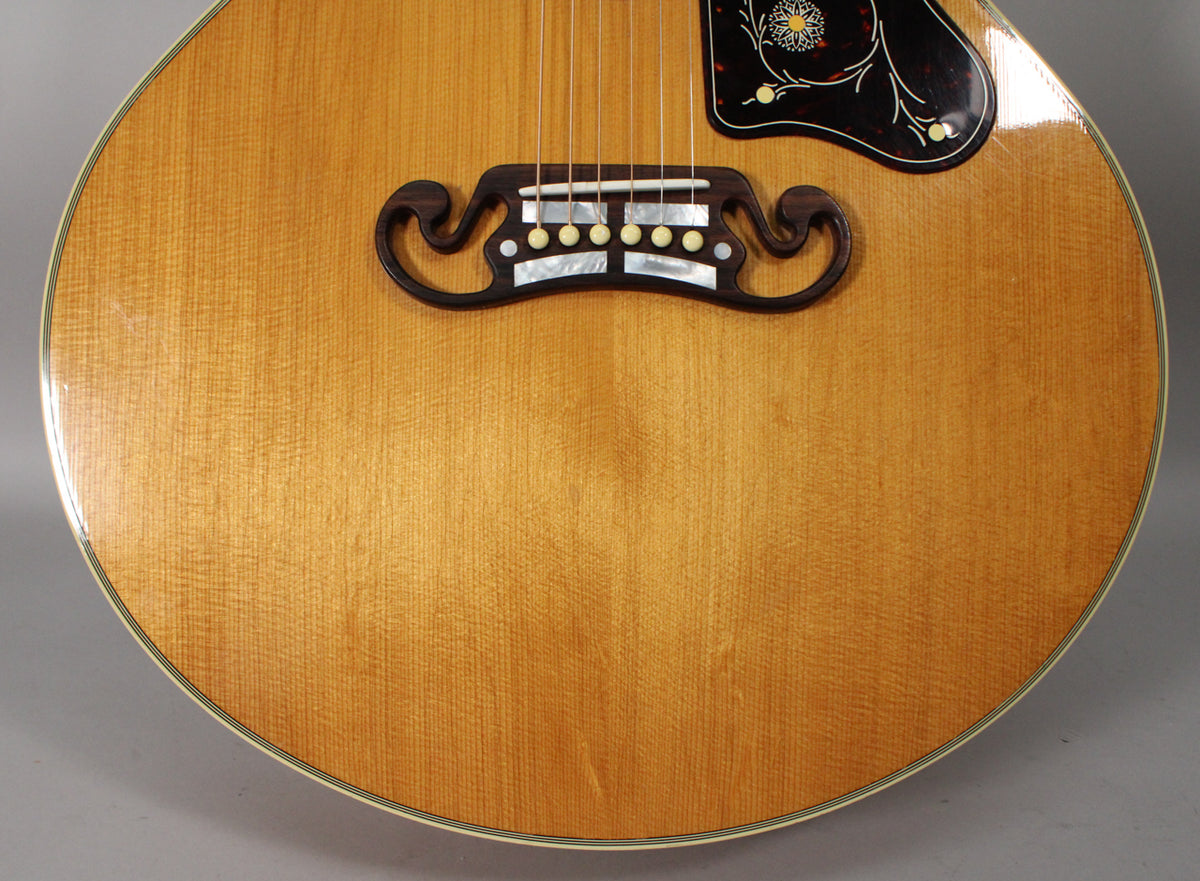 The natural finish acoustic guitar's lower bout is framed, showing its ornate dark wood bridge with mother-of-pearl inlay and light pin dots.