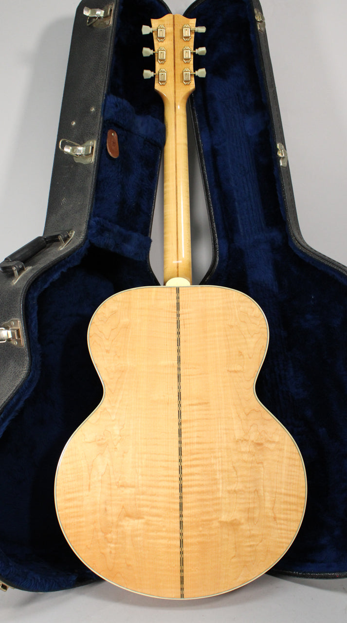 The back of a natural finish acoustic guitar with a flamed maple body and neck, resting in a dark blue plush-lined case.