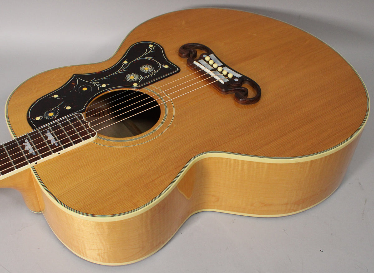 Angled view of a natural finish acoustic guitar, showing the upper bout, ornate black pickguard, and carved bridge.