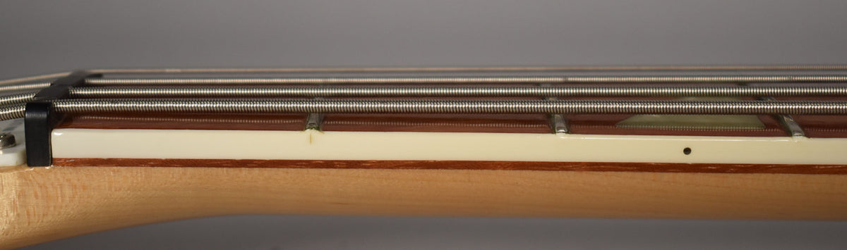 Close-up of the bass neck from the side, showing the light maple wood, cream binding, and a small chip near the nut.