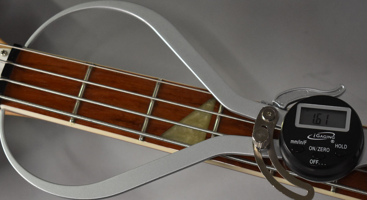 Close-up of the bass fretboard with a digital string action gauge showing 1.61mm, highlighting the light-colored sharkfin inlays.