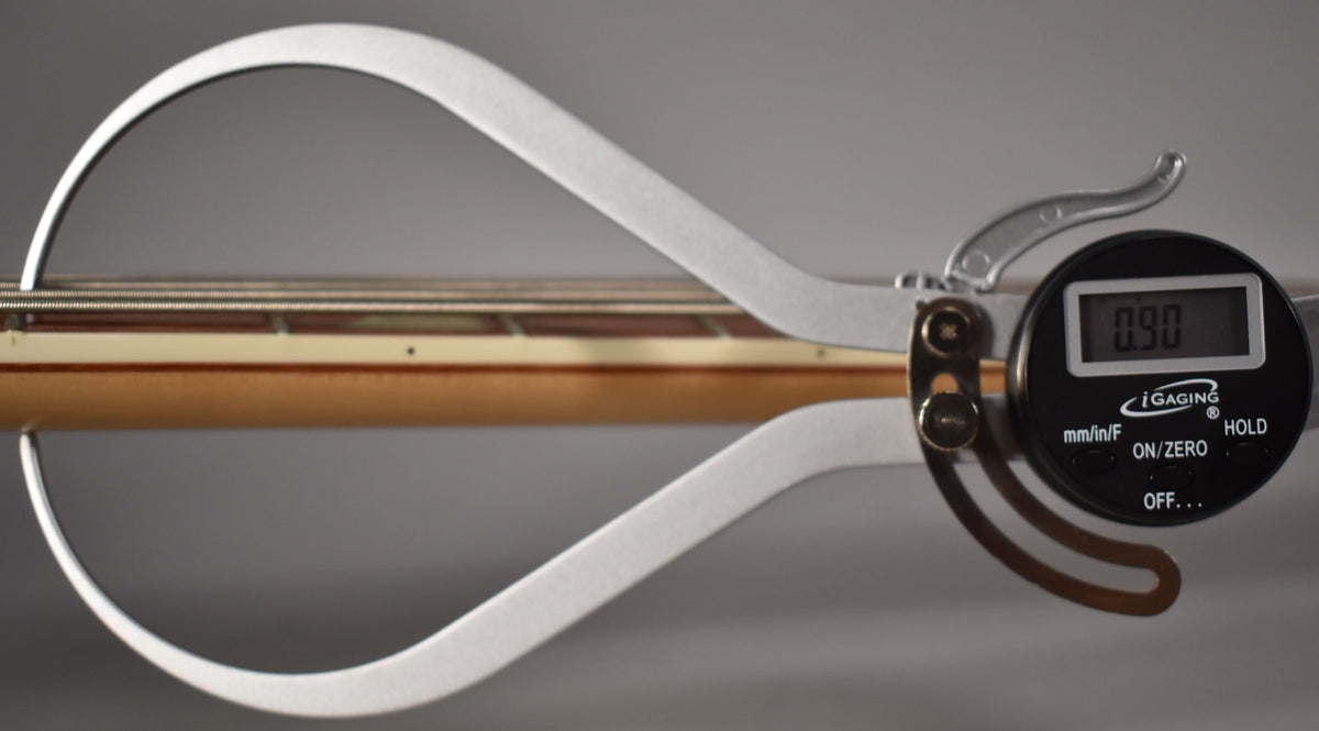 A digital caliper measures the bass's string height at the 12th fret, showing 0.90mm above the light maple fretboard.